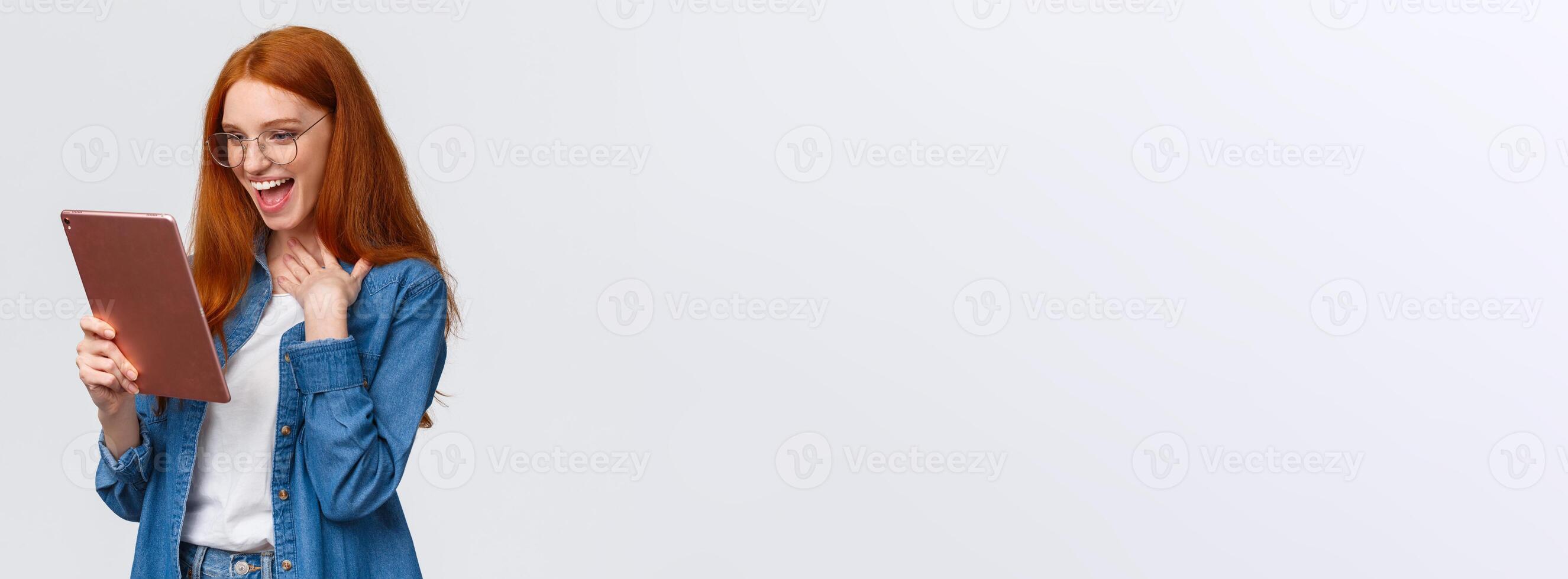 Excited, happy and surprised cheerful redhead woman messaging with friend using gadget, holding digital tablet, touch chest pleased and amazed, laughing out loud looking device screen photo