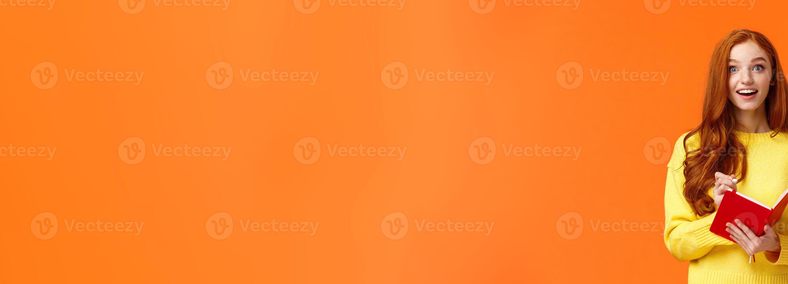 Inspired and upbeat, dreamy fascinated redhead girl writing down interesting lecture, smiling and gazing with admiration or enthusiasm, fill-in notebook schedule, standing orange background photo