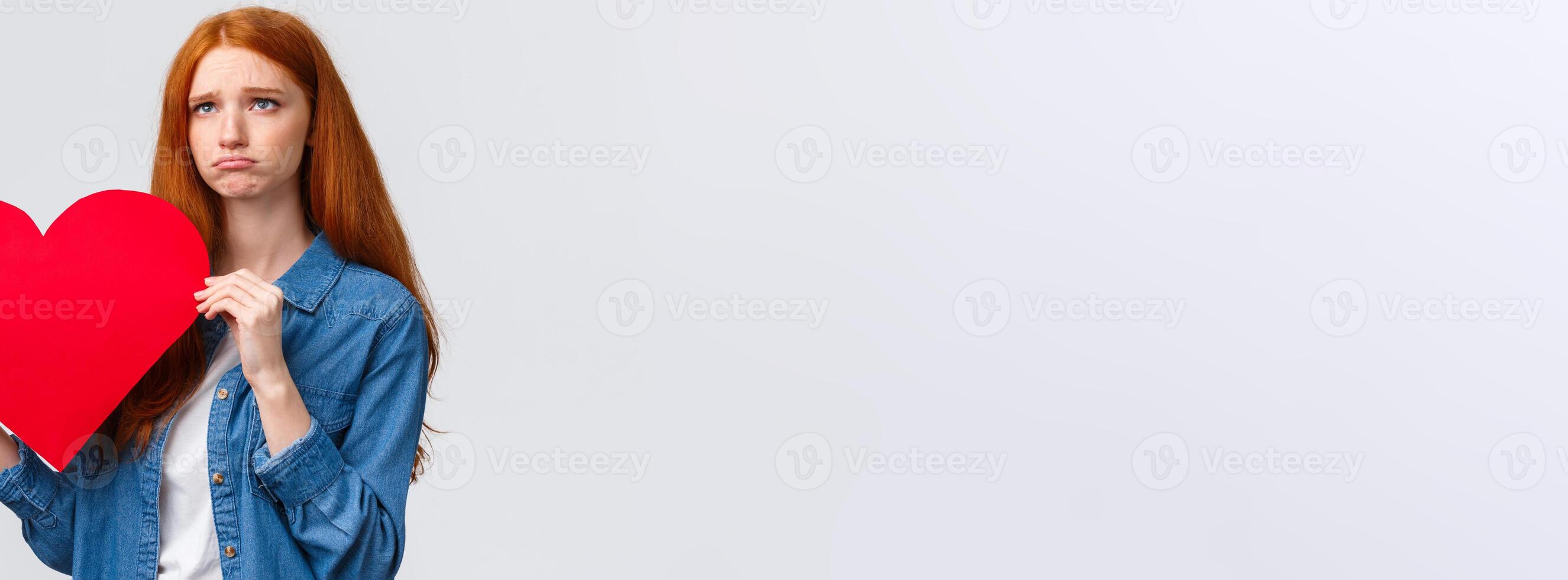Unhapy brokenhearted cute redhead girl, sulking and looking up uneasy, distressed holding big red valentines day heart, was rejected, showed her feelings standing white background photo