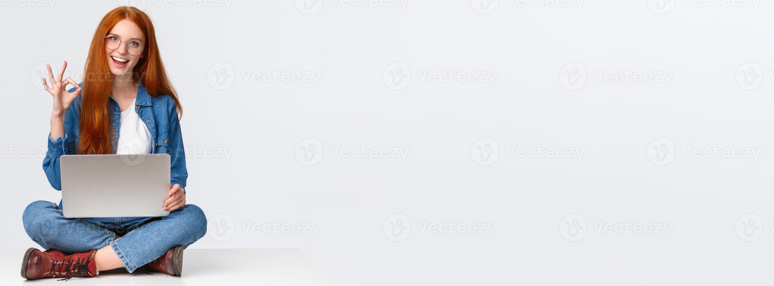 Dont worry, all done, everything under control. Confident happy and relaxed redhead girl saying all okay, show ok gesture while sit on floor with legs crossed, holding laptop, white background photo