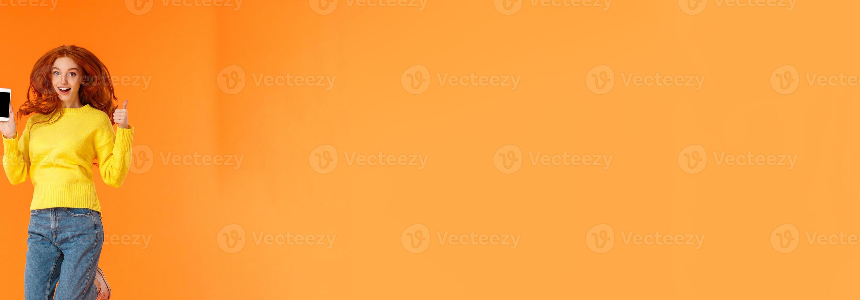 Advertising, technology and holidays concept. Vertical full-length shot cheerful gorgeous ginger girl, redhead woman jumping over orange background with smartphone, show thumb-up photo