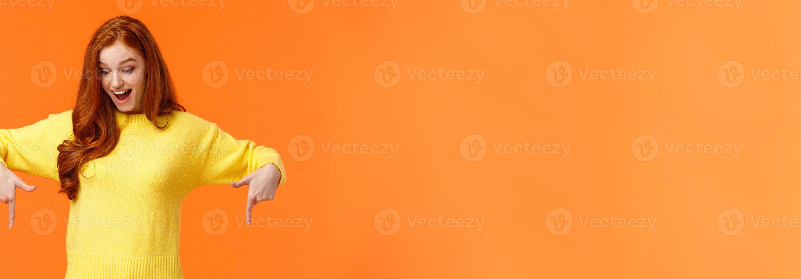 Wondered and excited, amused cute redhead woman in yellow sweater pointing fingers down, look bottom and smiling, check-out product promo, winter holiday event, orange background photo