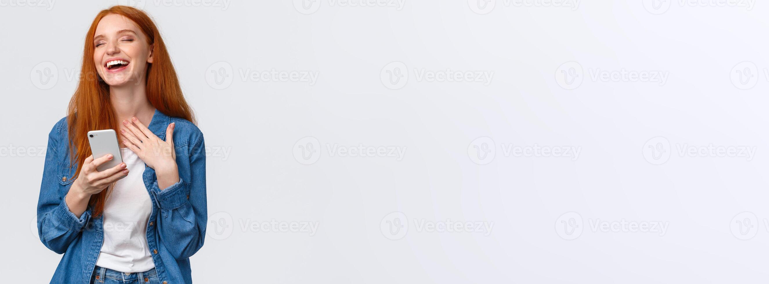 Waist-up portrait carefree and joyful, alluring redhead caucasian girl receive funny meme message, holding smartphone, laughing and close eyes dreamy, standing white background photo