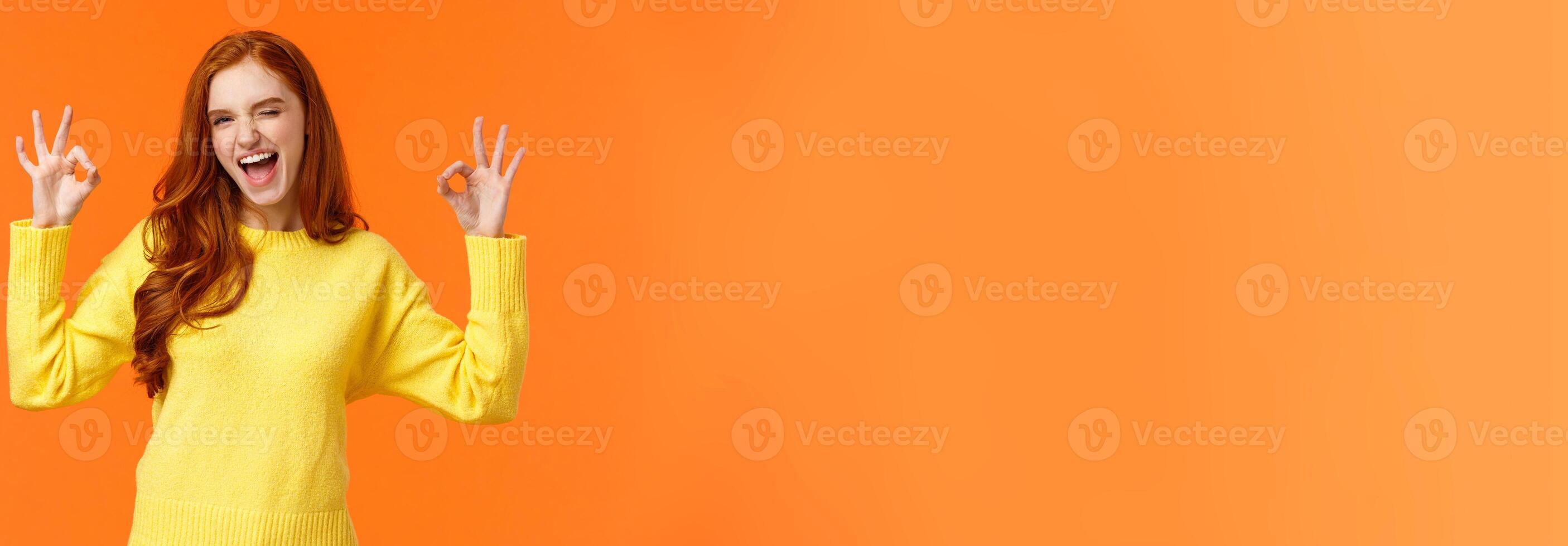 Cheerful and positive, enthusiastic redhead female in yellow sweater winking in approval, smiling joyful, give permission, positive reply, rate good movie, nod agreement, recommend, orange background photo