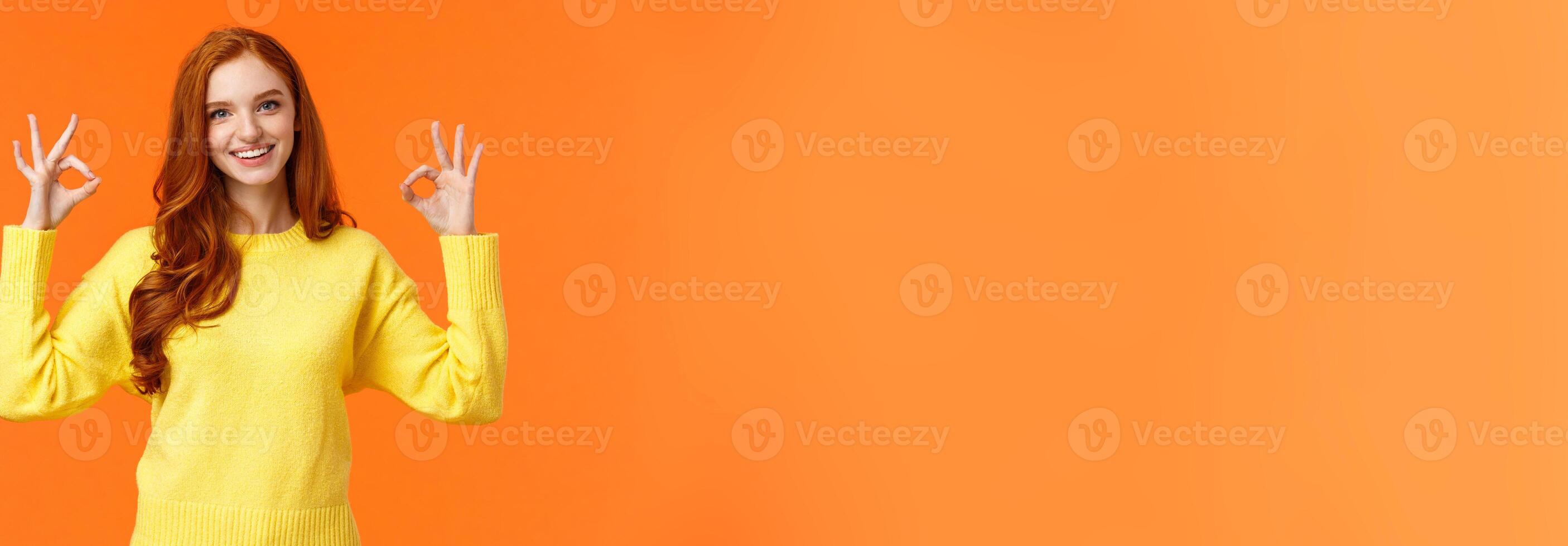 Perfect idea concept, acception. Good-looking redhead girl in yellow sweater, showing okay, good or approval sign smiling nod agreement, satisfied with result, all perfect, orange background photo