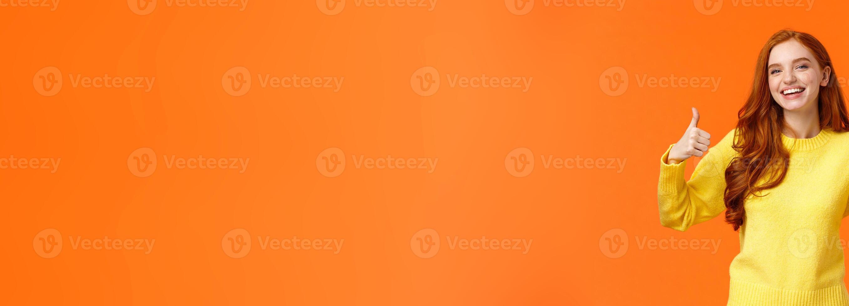 Holidays, gestures, people concept. Cheerful cute redhead girl smiling and showing thumbs-up recommend product, give positive reply, agree or like something, nod agreement, orange background photo