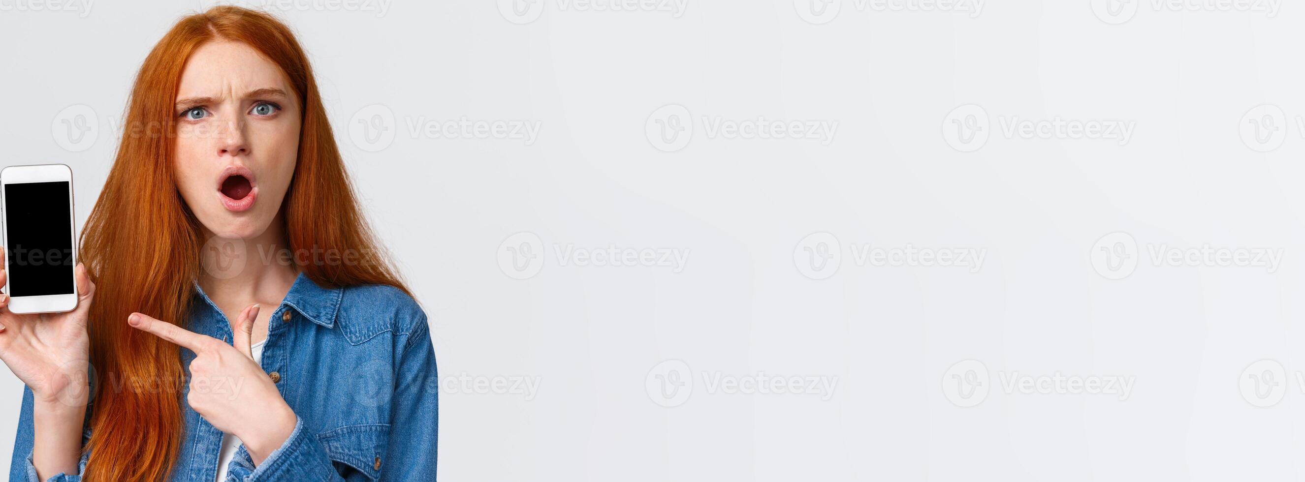 Close-up portrait angry and pissed-off redhead girl showing photos from social media ex-boyfriend complaining to friend, pointing finger smartphone display and frowning upset, white background
