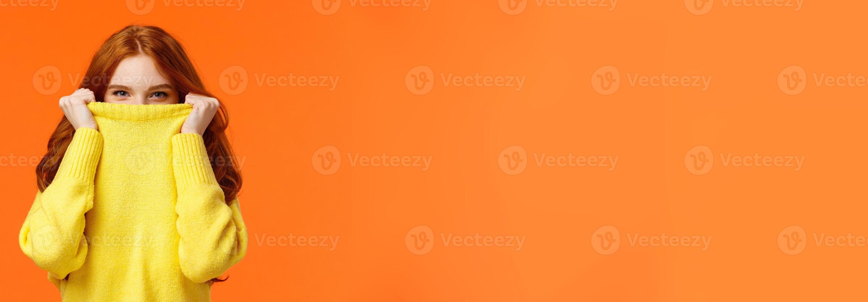 Girl hiding from winter cold in her warm soft sweater. Cute redhead woman pulling collar on nose and peeking at camera with smiling eyes, glancing at you happy, orange background photo