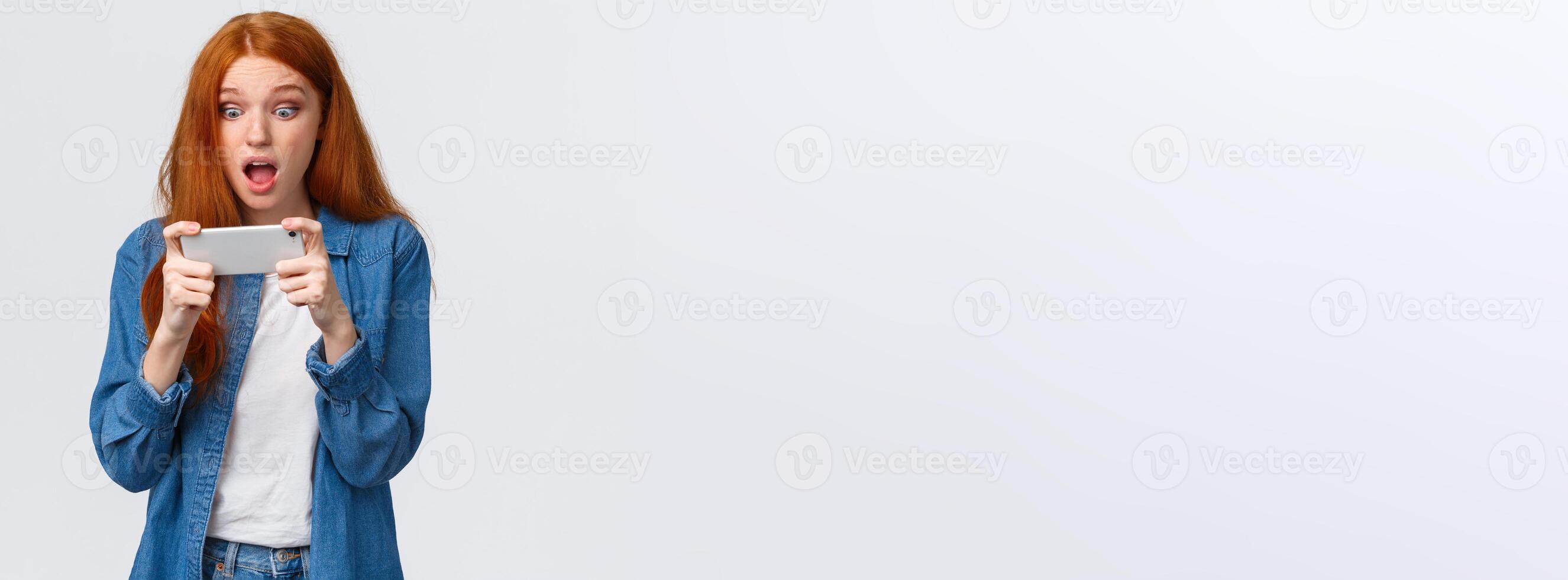 Astonished startled cute redhead woman just won challenge in awesome game, holding smartphone horizonally, playing racing, stare display amazed, cant belive she lucky, white background photo