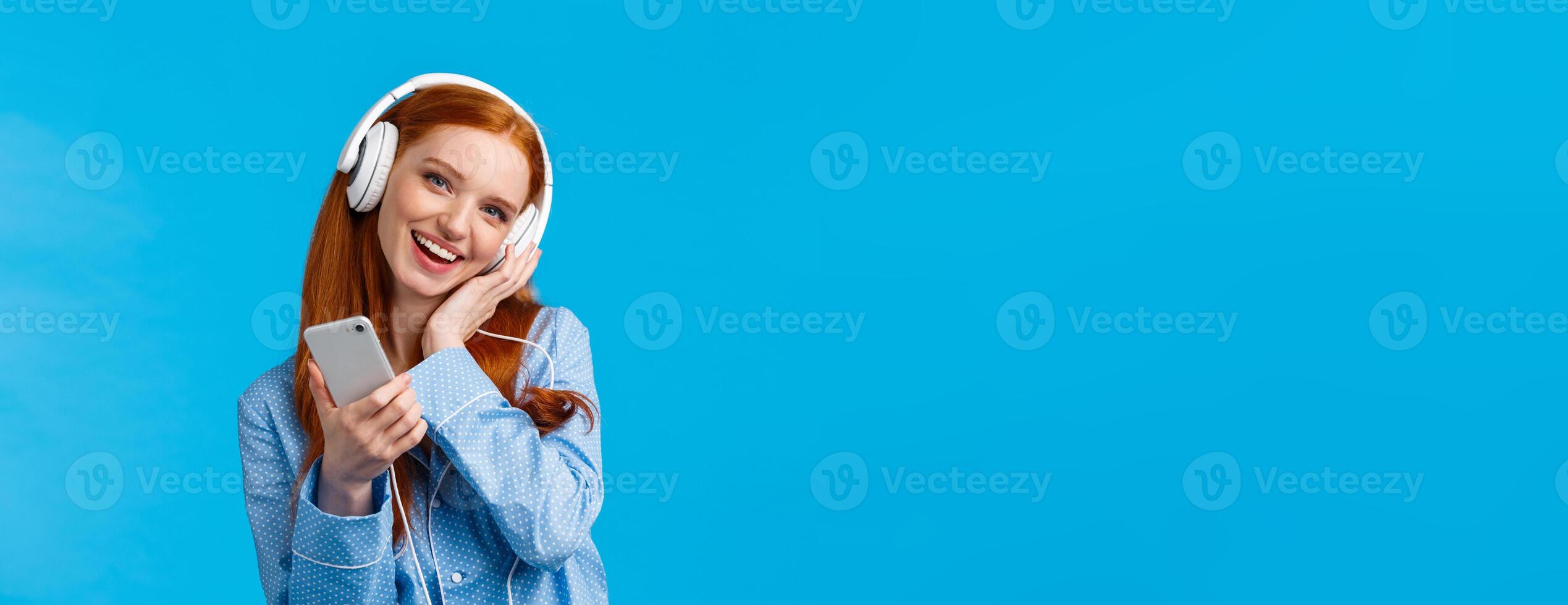 Lovely young dreamy girl fantasizing as listening favorite songs, voice of singer, tilt head and touching headphone as enjoying music earphones, holding smartphone and smiling, blue background photo