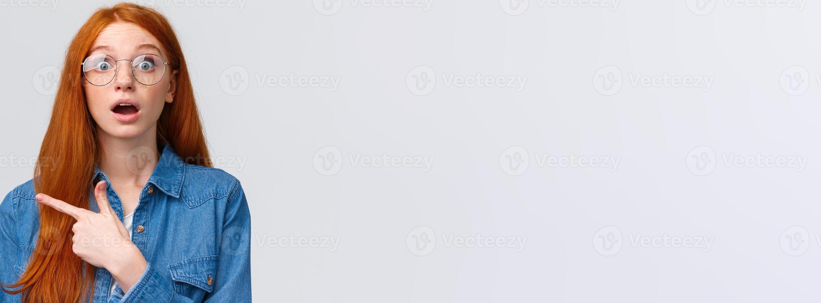 Surprised and wondered, amused attractive redhead woman in glasses, red natural hair, freckles, blue eyes open mouth fascinated and curious, pointing finger left, asking question about banner photo