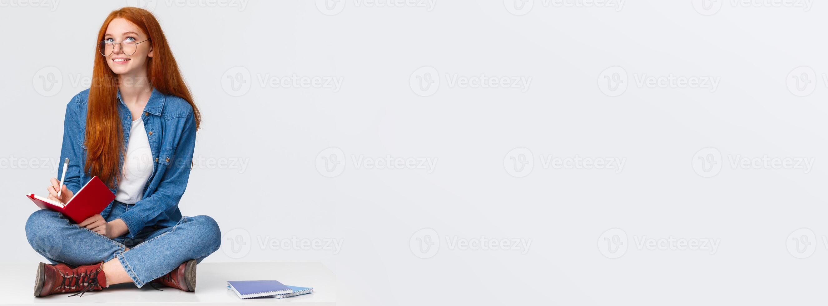 Dreamy and creative cute diligent female student with red hair, in glasses, sitting on floor with legs crossed surrounded by notebooks writing something, creating poem, look up thoughtful photo