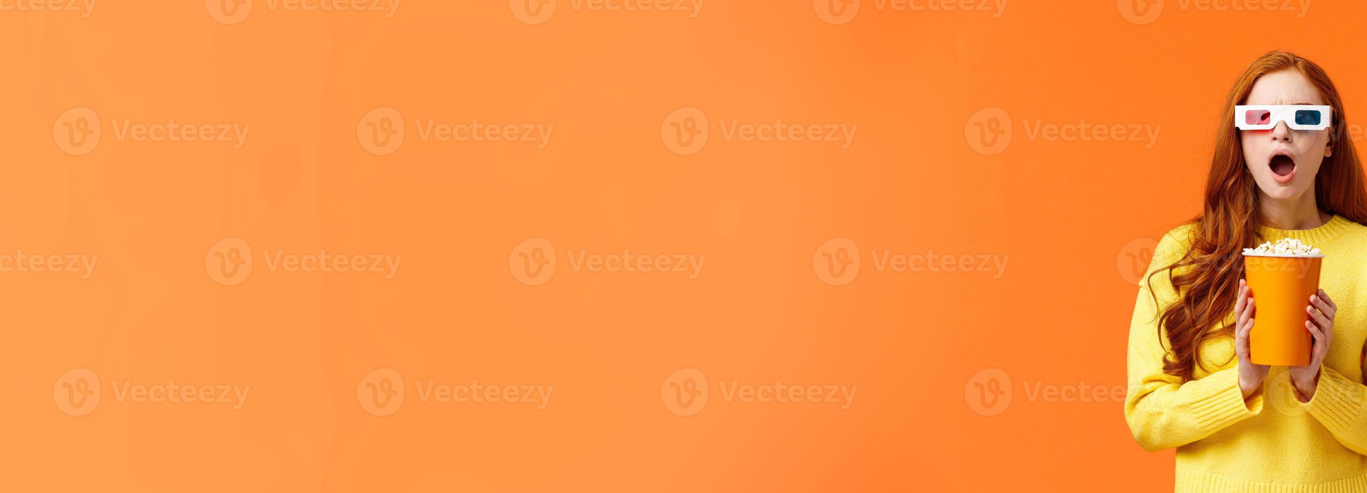 Shocked and tensed redhead woman seeing intense fight on screen as watching movie at cinema, wear 3d glasses, holding popcron, frowning and gasping concerned, orange background photo