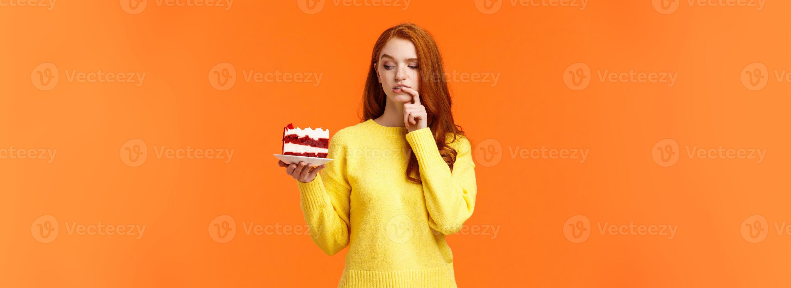 Diet, healthy lifestyle and junk food concept. Cute redhead woman cant resist temptation bite tasty cake, holding dessert on plate and biting lip thinking how many calories in it, orange background photo
