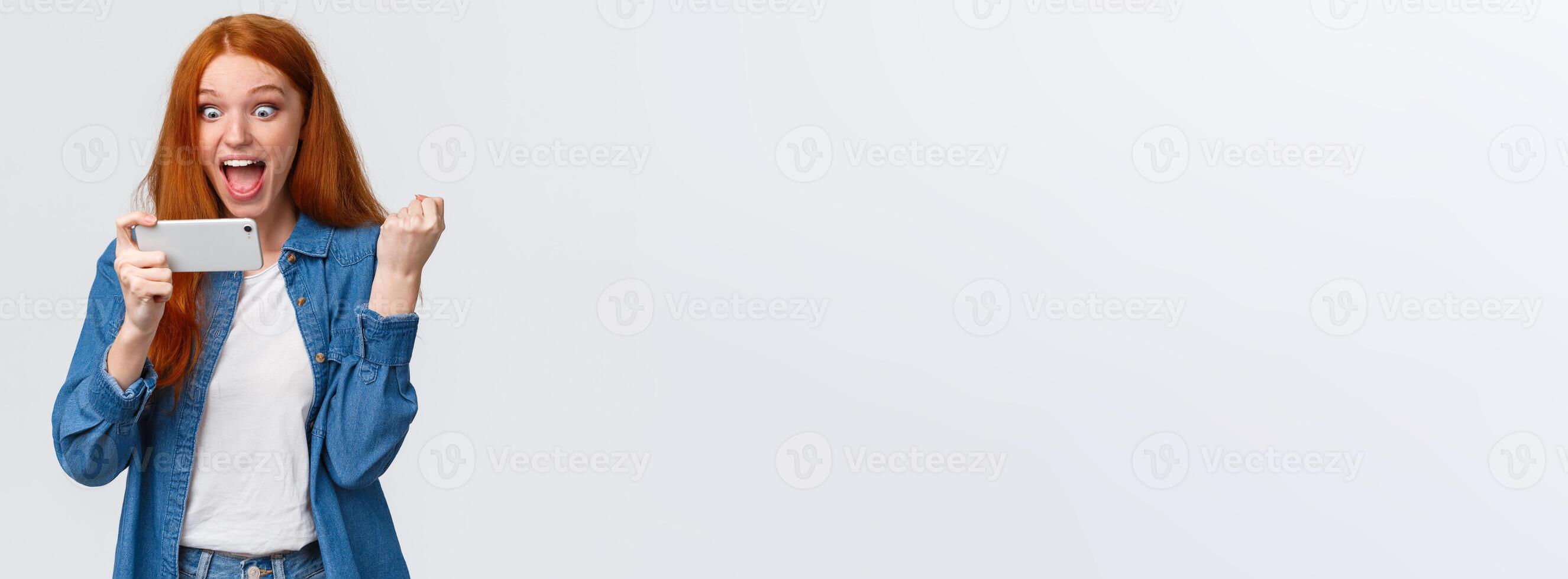 Achievement, goal and success concept. Cheerful happy cute redhead teenage girl fist pump triumphing, staring at smartphone display astonished and satisfied, winning game challenge photo
