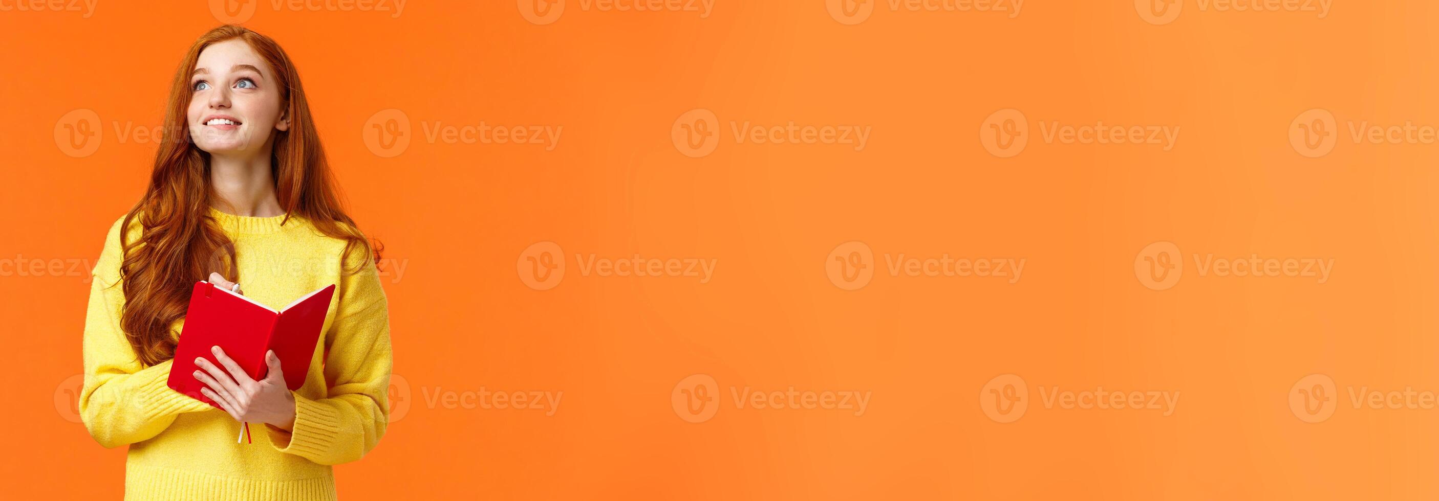 Dreamy, inspirational cute redhead female student writing daily tasks, to-do list in red lovely planner, look upper left corner thoughtful as smiling, getting inspiration to write novel, orange wall photo