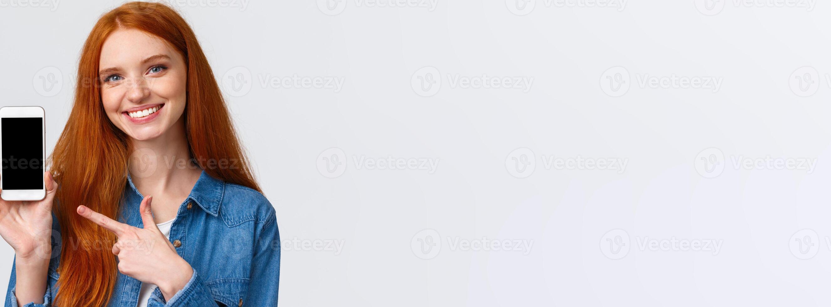 Girl found her date on this app. Cheerful attractive redhead woman with blue eyes give advice, showing smartphone display and pointing finger mobile screen, smiling give recommendation photo