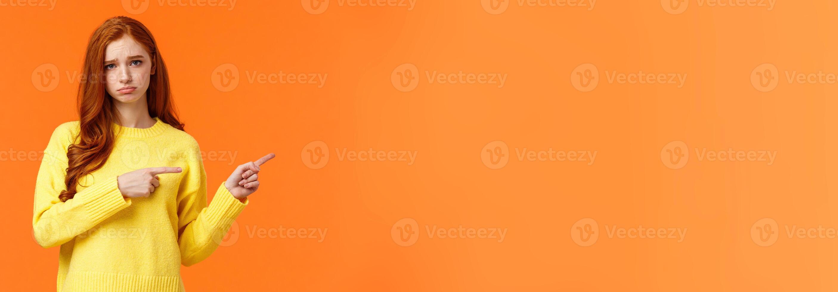 Sulking moody and sad cute redhead girlfriend pointing right and frowning, standing unhappy with bad mood, envy someone, begging boyfriend book tickets for winter ski-resort, orange background photo