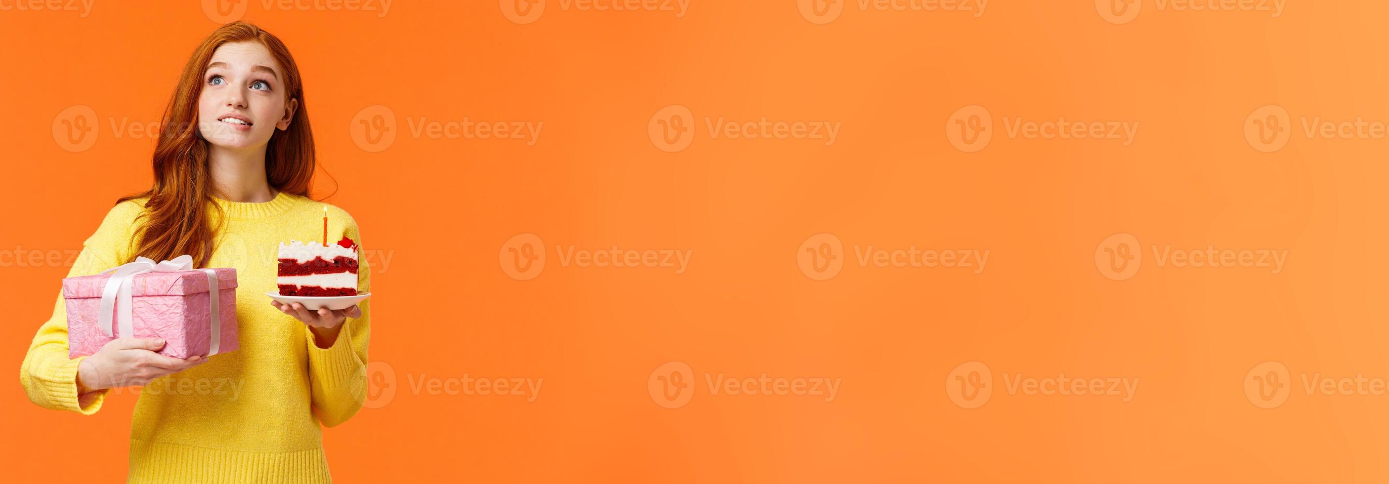 Dreamy, silly cute redhead girl with passionate, admiring look, biting lip have desire or wish, holding piece b-day cake delicious dessert, blowing candle, receive birthday gift in box, orange wall photo