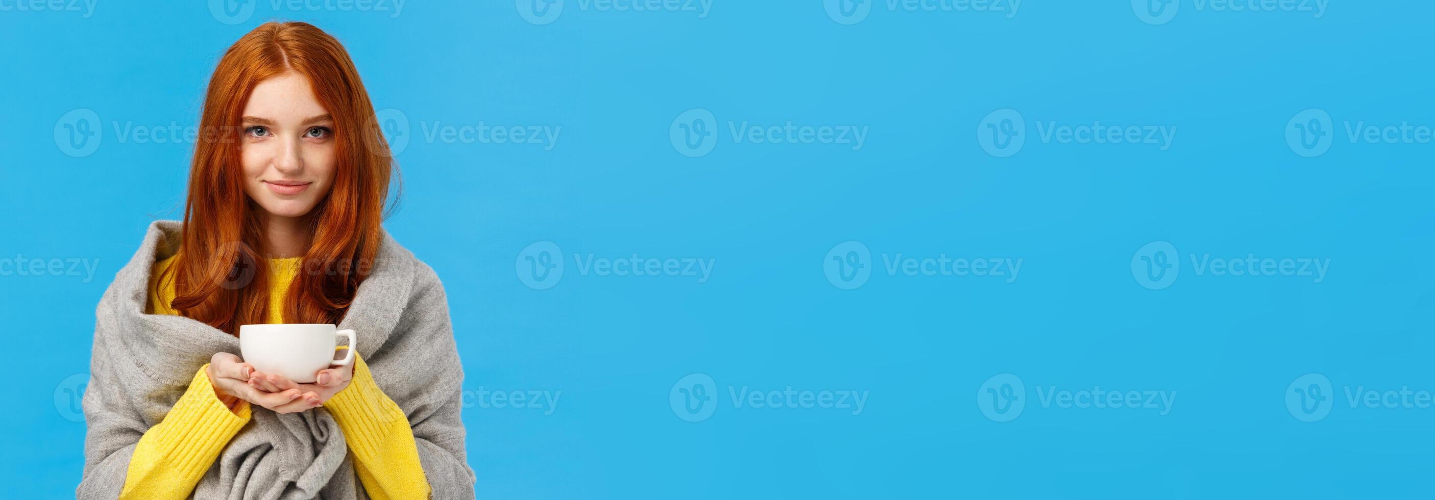 Redhead girl trying get warm, feeling cold wrapping herself with scarf on shoulders, drinking hot tea, holding teapot and smiling lovely, standing tender and cute blue background photo