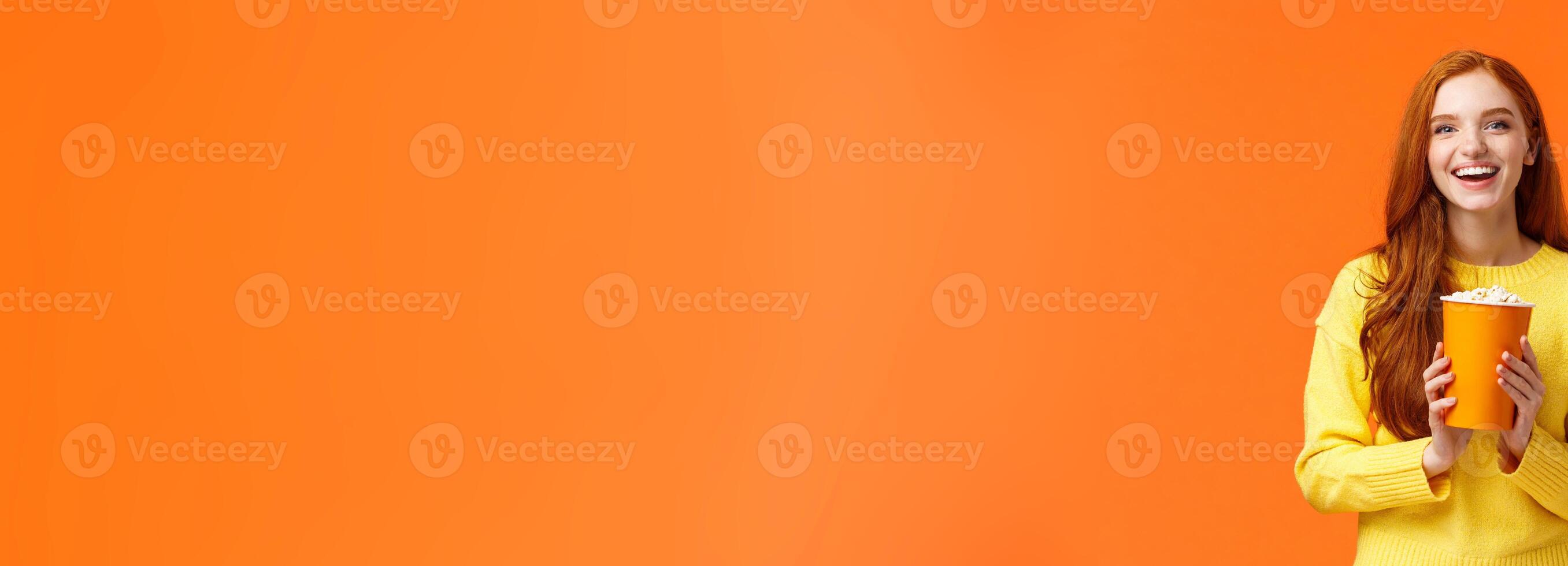 Cheerful cute redhead curly girlfriend holding popcorn chat with boyfriend before movie start at cinema, smiling joyfully, like attend premieres in theatres, standing overjoy orange background photo