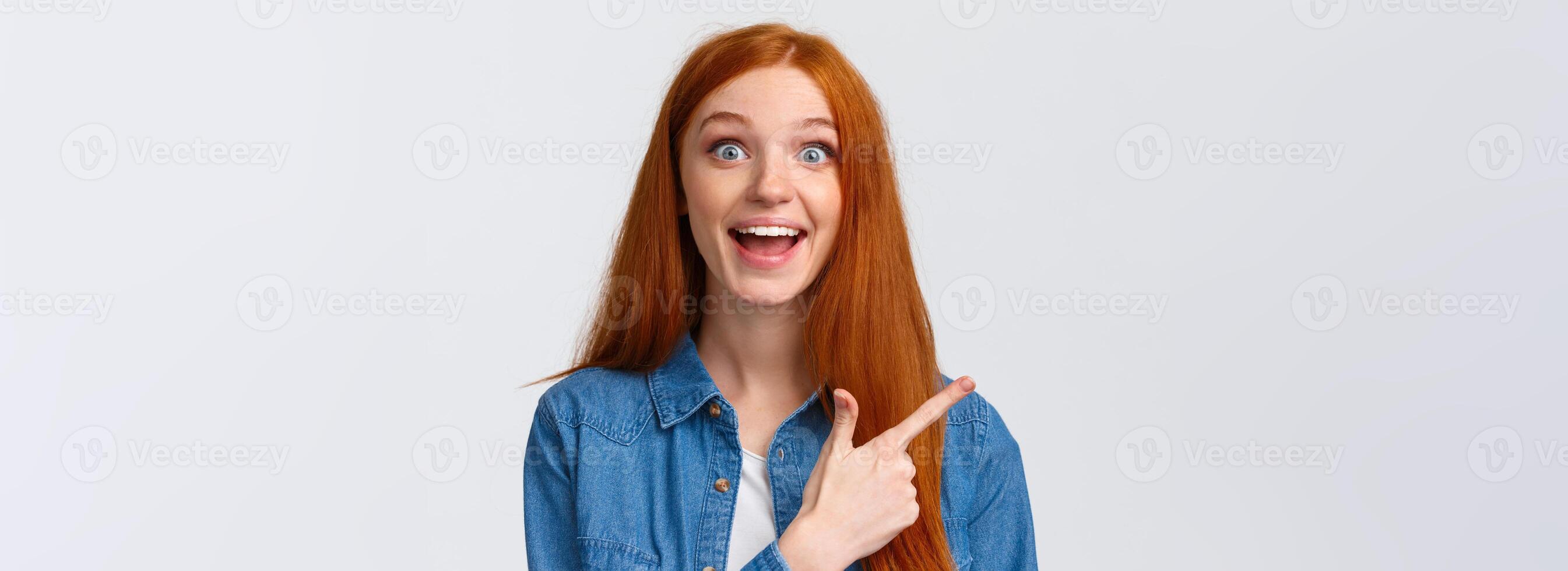 Close-up portrait excited happy ginger girl, foxy hair and blue eyes, smiling upbeat with amazement and joy, pointing finger left, attend amusing cool party, telling about it, white background photo
