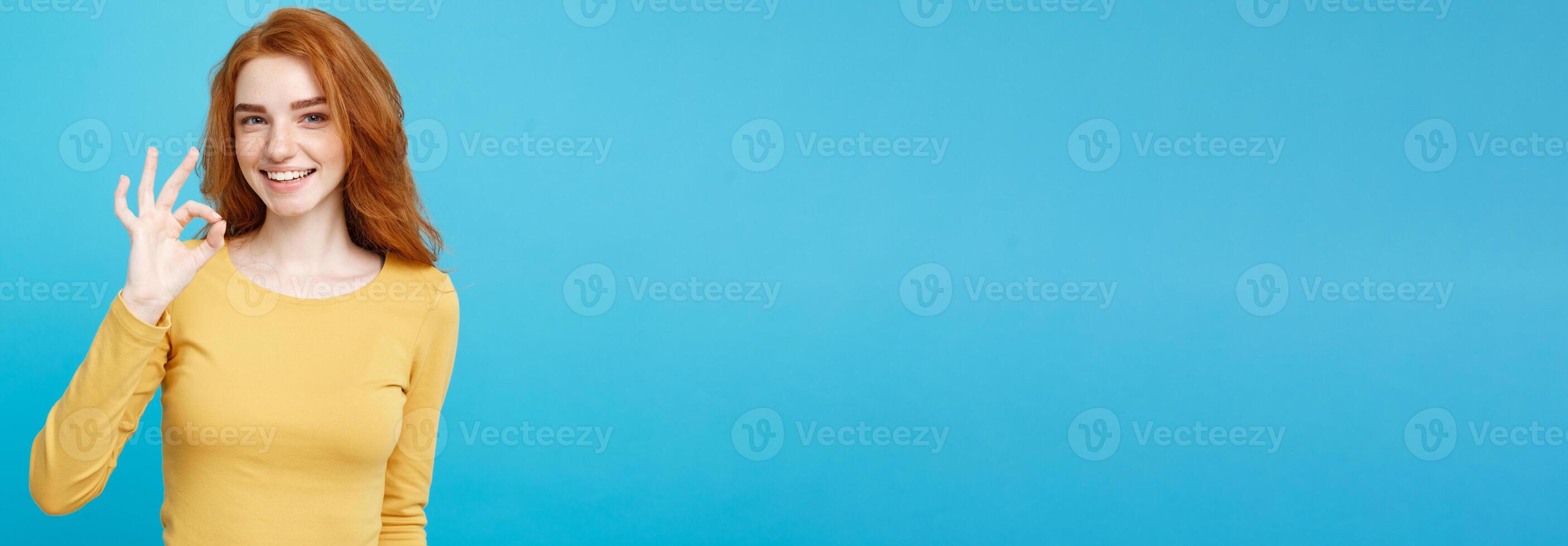 Fun and People Concept - Headshot Portrait of charming ginger red hair girl with freckles smiling and making ok sign with finger. Pastel blue background. Copy Space. photo