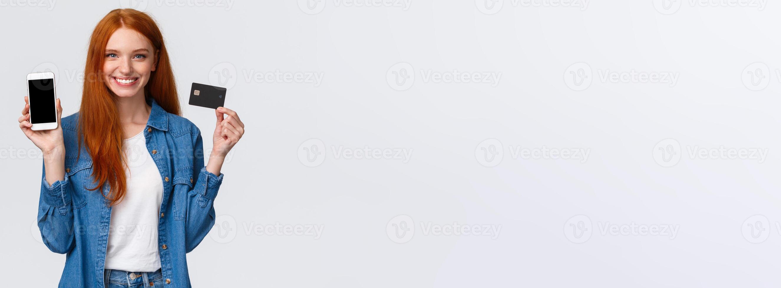 Waist-up portrait confident female with red long hair, blue eyes, holding credit card and showing smartphone display, easy banking service, payment for bills internet, smiling satisfied photo
