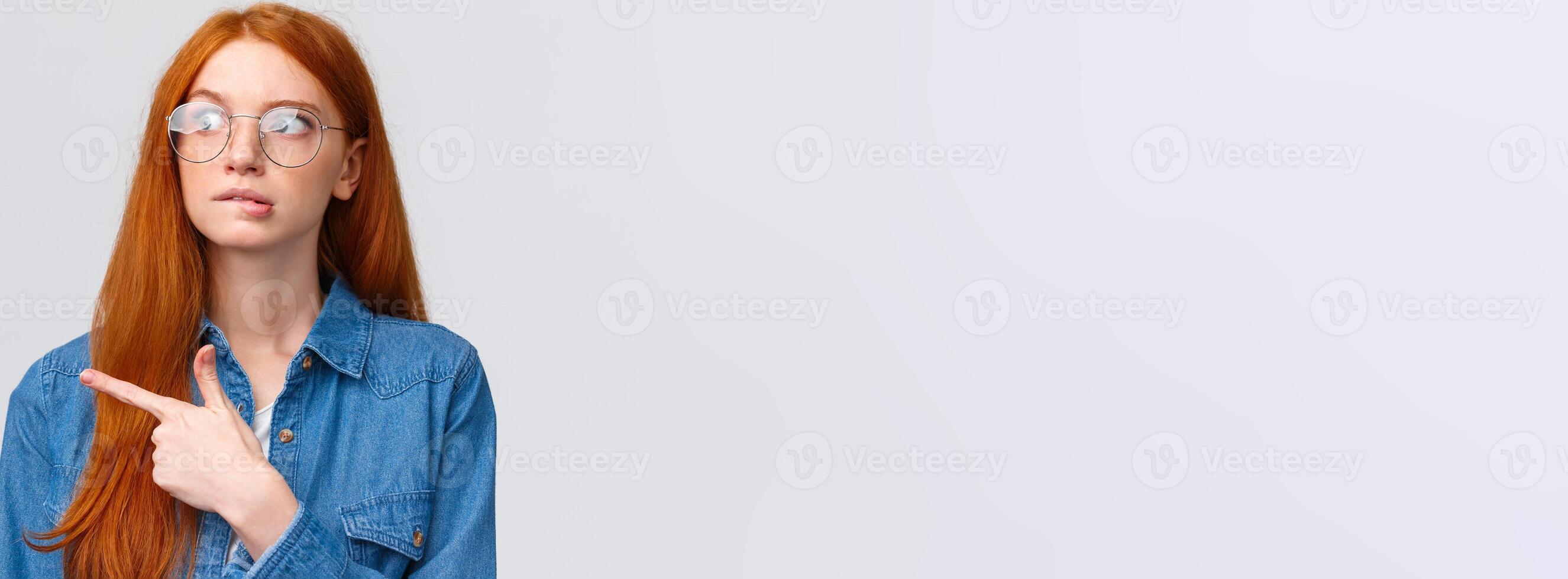 Unsure smart and cute redhead teenager in glasses, female student biting lip curious looking and pointing left, thinking, deciding what pick, making choice, standing over white background photo