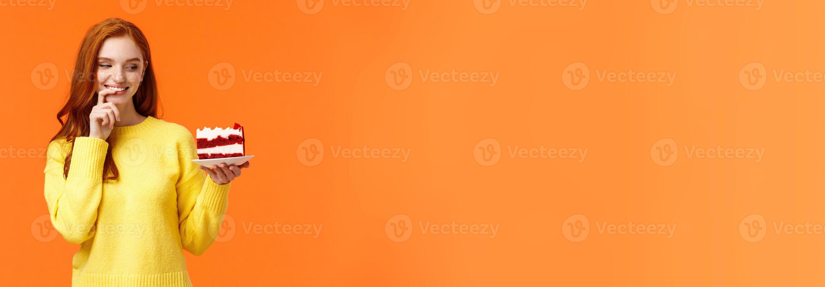 Cheerful and satisfied, happy redhead woman have cheat-day eating delicious food, holding tasty piece cake, biting lip and smiling, cant resist temptation, desire take bite, orange background photo