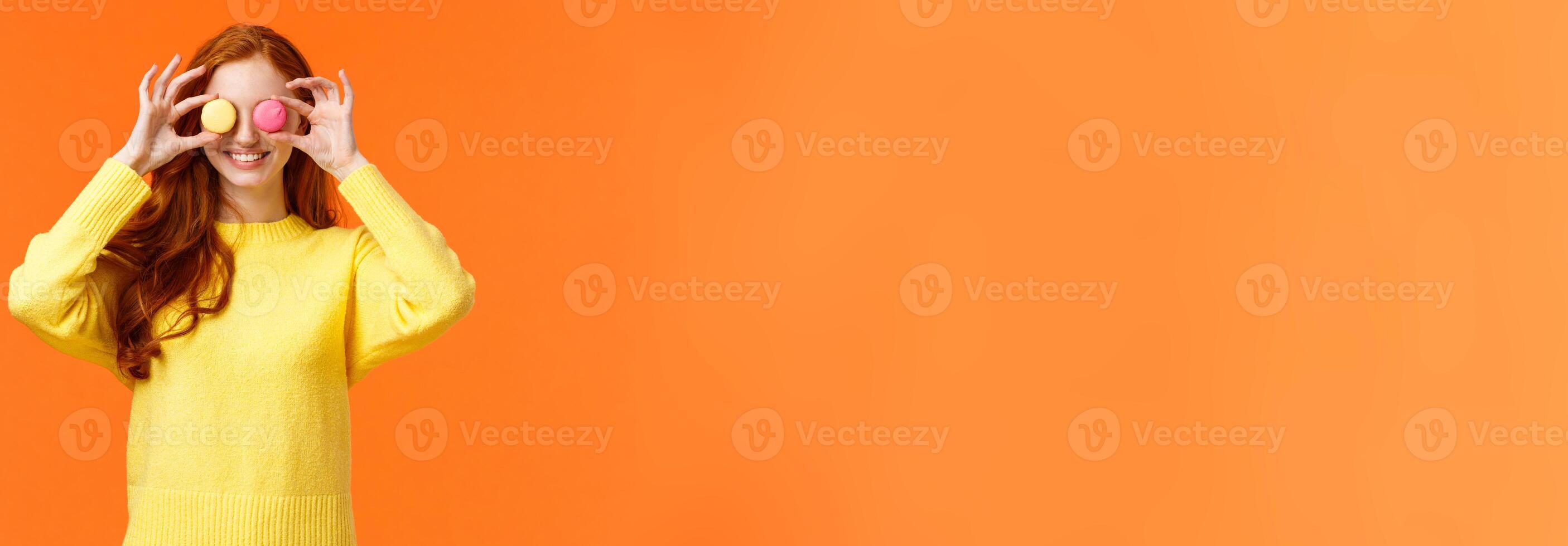 Fashion, food and consumer concept. Cheerful silly redhead woman fooling around, playing with dessert, make eyes from macarons and smiling joyfully, standing orange background photo