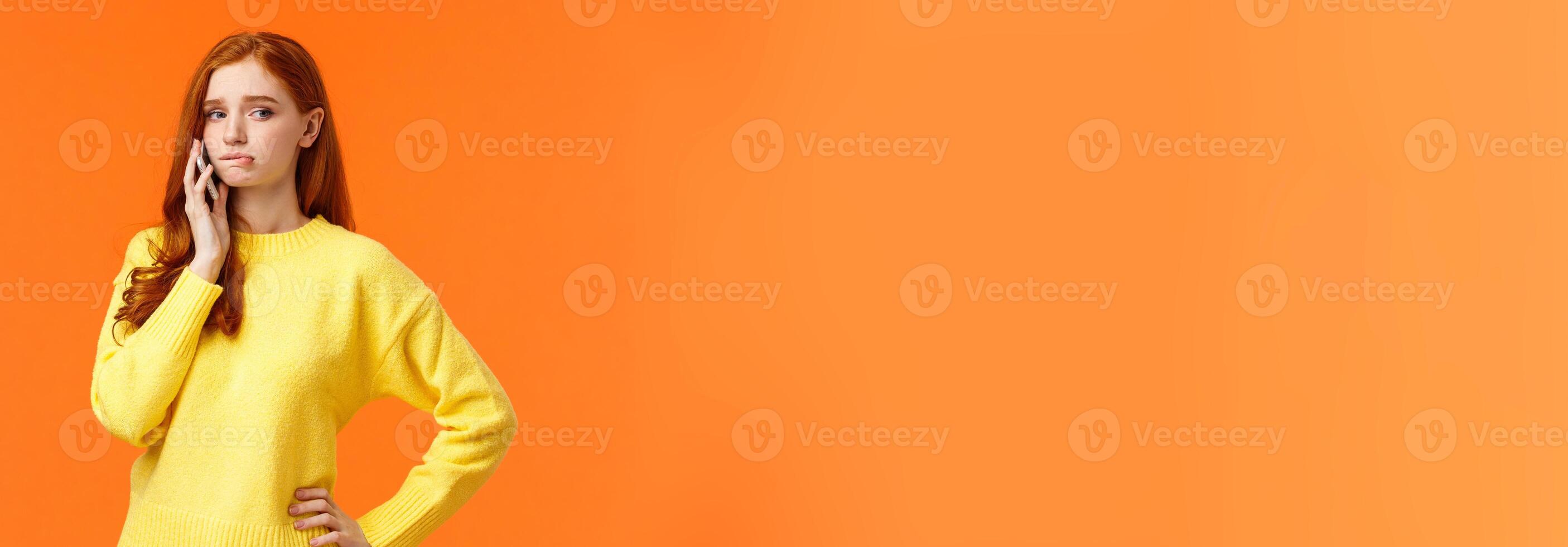 Girl catching anxiety calling someone. Perplexed or nervous cute redhead woman biting lip and looking away as having conversation on mobile phone, standing orange background sad photo
