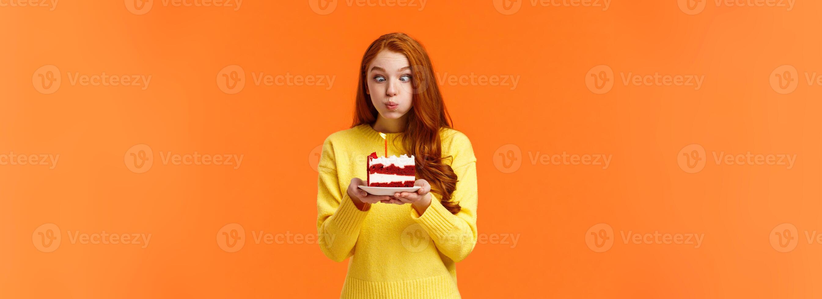 Funny and silly cute redhead b-day girl squinting eyes making goofy expression, pouting as holding birthday cake and blowing-out candle to make wish, having fun at party celebrating, orange wall photo