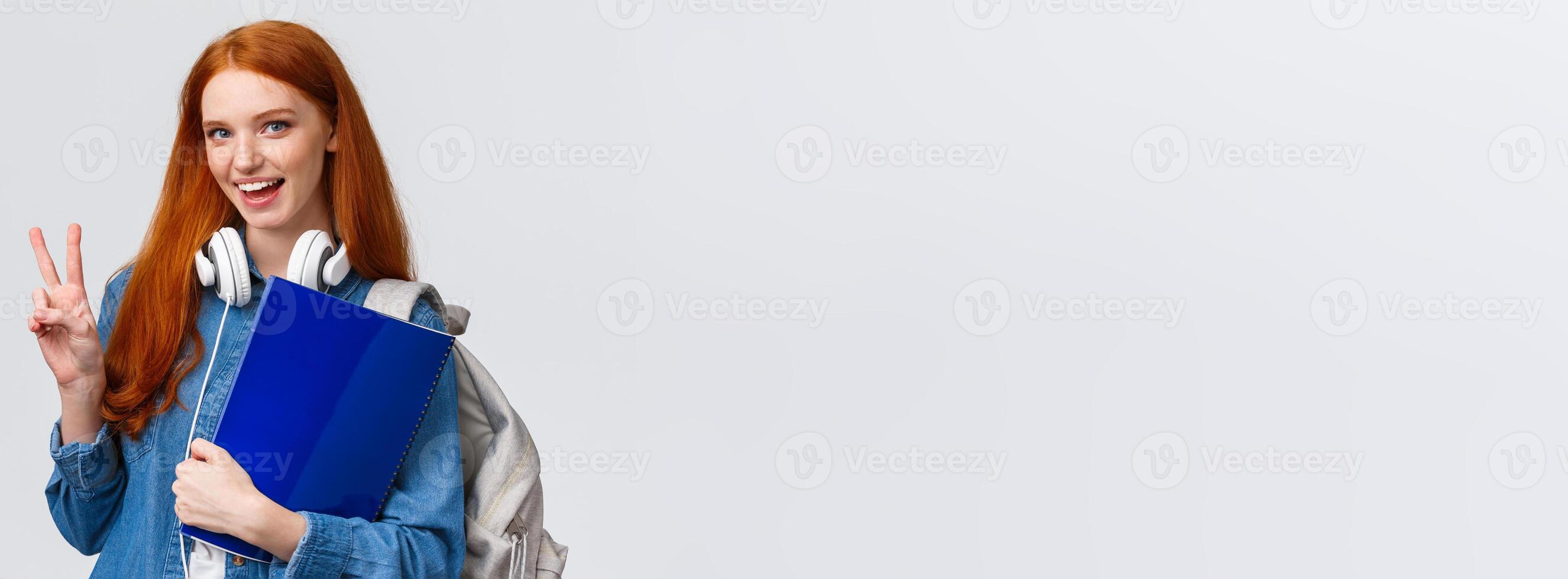 Lovely friendly college student, redhead girl with papers, documents for school, holding backpack and wear headphones over neck, make peace gesture informal greeting classmate, white background photo