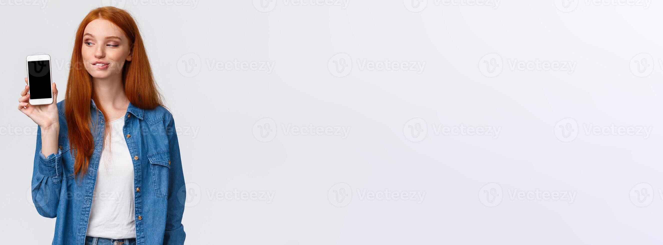 Curious and daring sassy redhead girl tempting buy something in online store, have interesting plan, smirk and peek at smartphone screen with pleased expression, standing intrigued white background photo