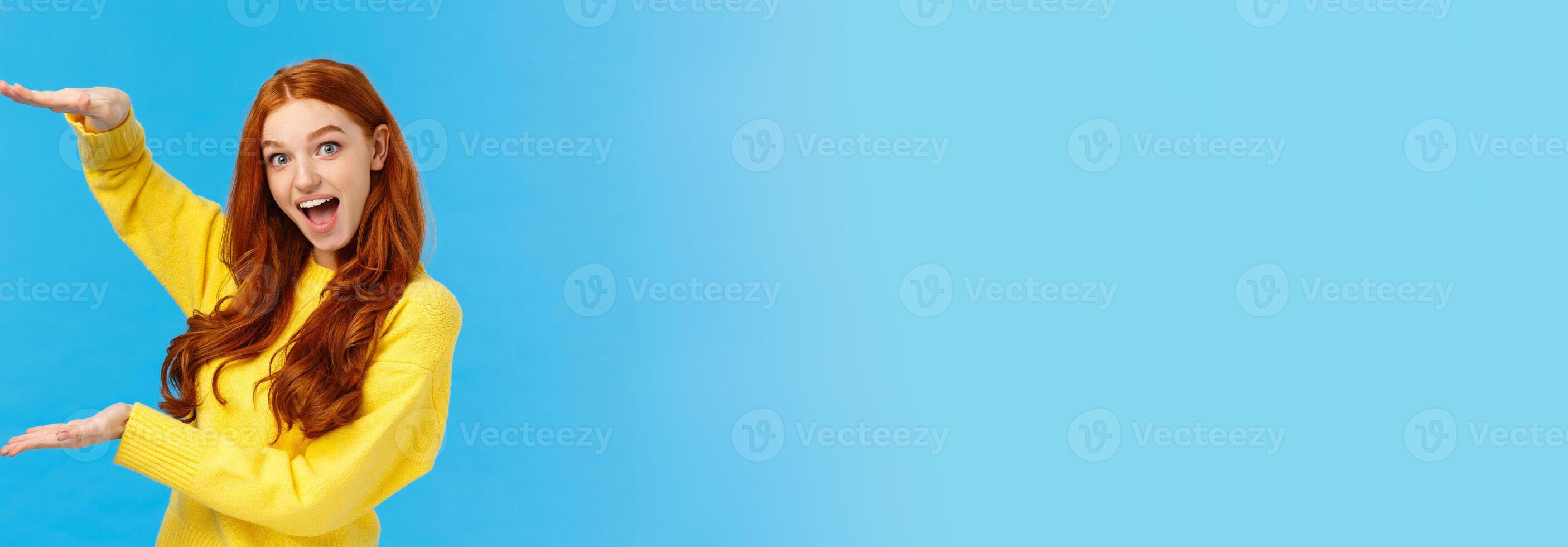 Excited lively good-looking redhead ecstatic girl smiling fascinated and amused, showing big, large object, shaping size with raised hands over blue background, standing joyful, impressed photo