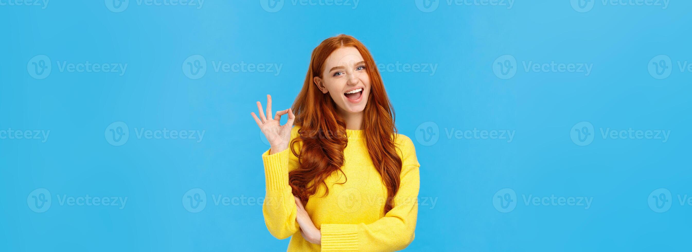 Waist-up shot carefree cheerful and friendly redhead gorgeous female freelancer finished IT school and showing okay gesture, no problem sign, smiling unbothered, assure all good, blue background photo