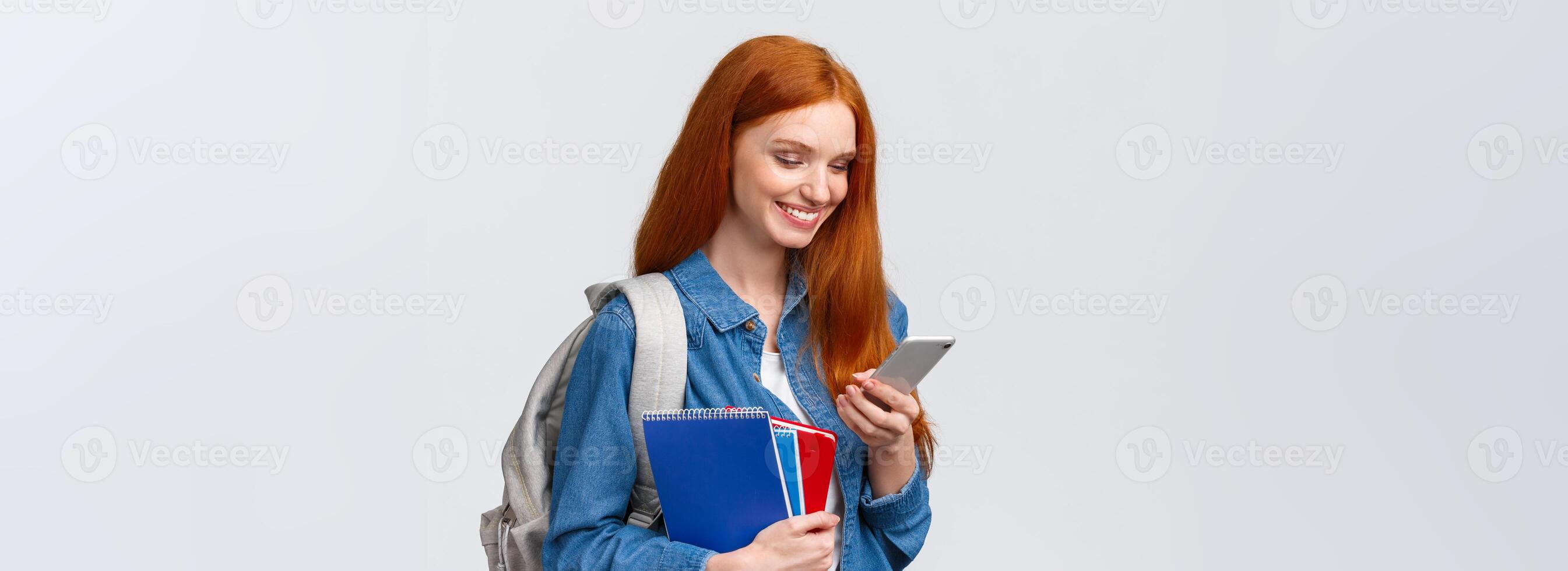 Lovely good-looking female with red hair, heading to college, holding backpack, notebooks smiling as answer friend message, chatting glancing mobile screen delighted, white background photo