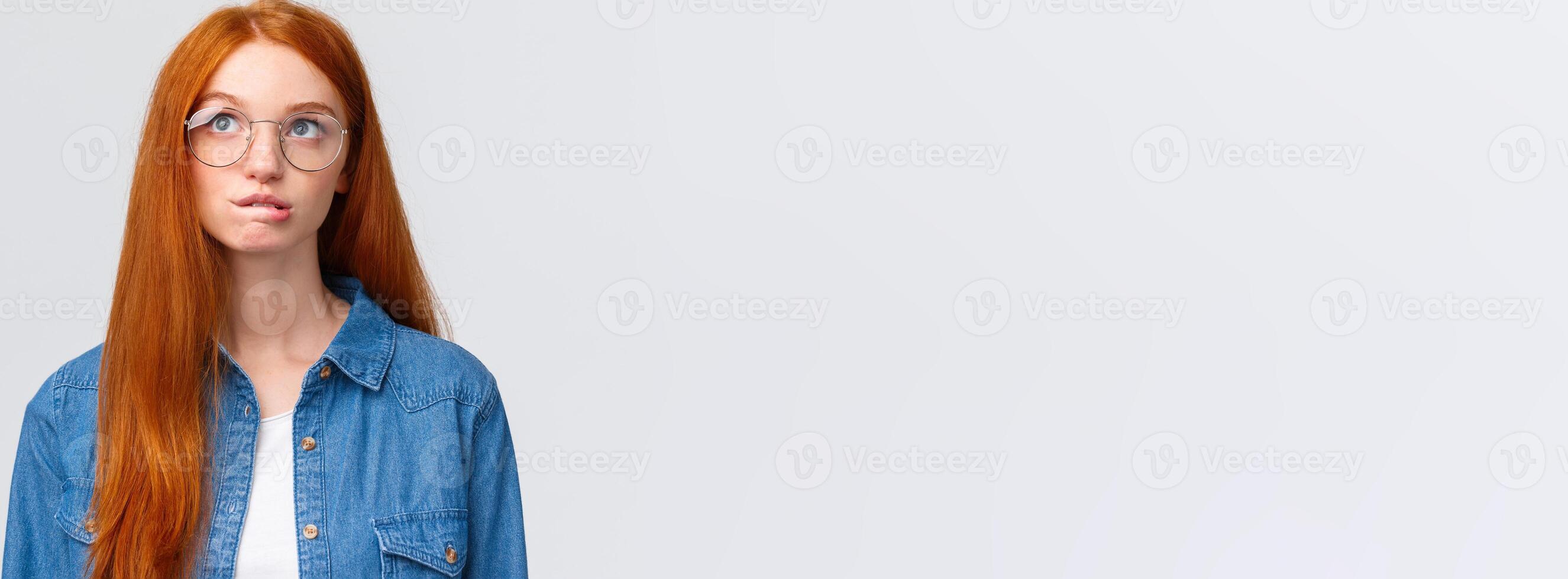 Thoughtful and curious cute redhead teenage girl, thinking, biting lip intrigued and looking up as planning something, recall, pondering between choices, standing white background photo
