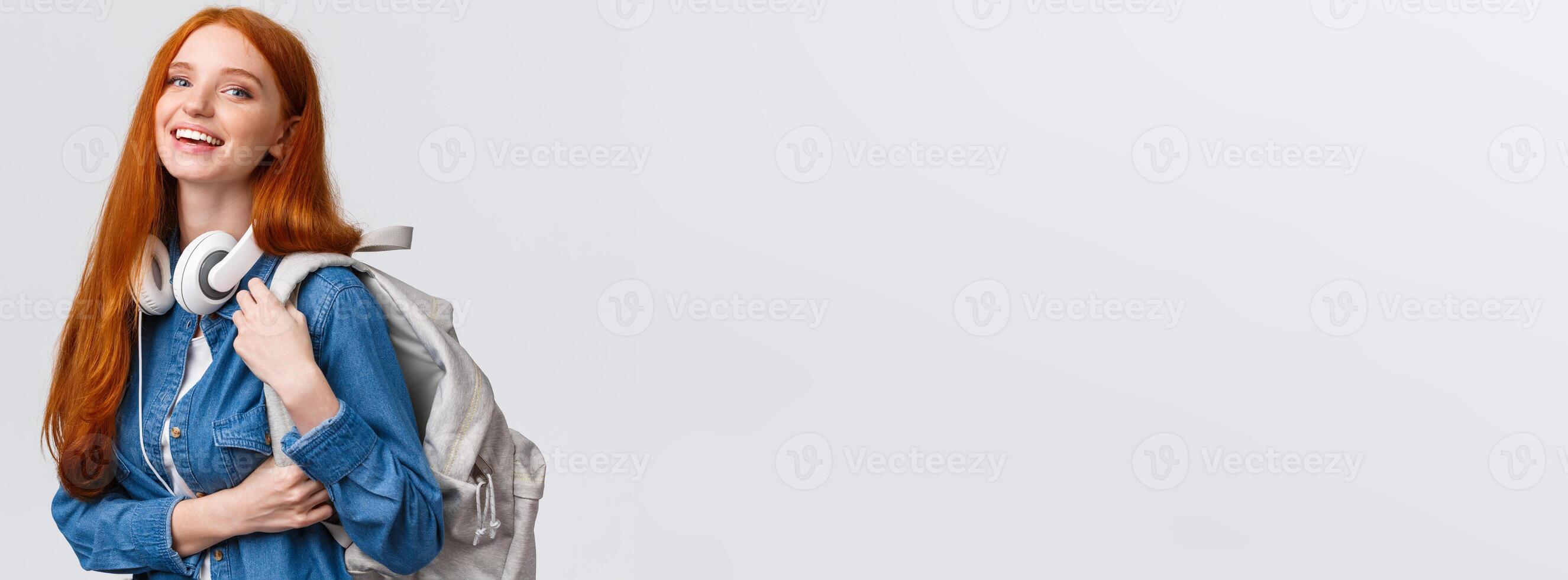 College life, modern lifestyle and education concept. Cheerful good-looking redhead female student with foxy long hair, wearing headphones over neck, backpack, smiling camera photo