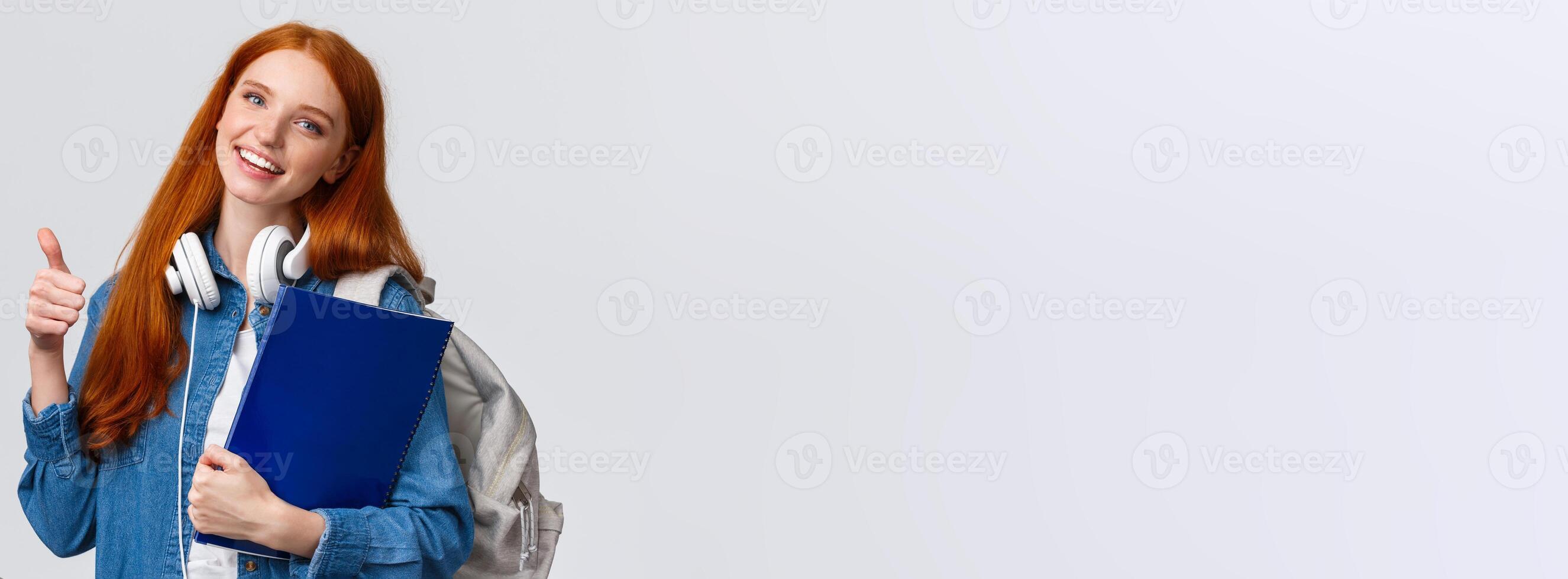 Education, teenagers and student lifestyle concept. Cheerful redhead lovely girl with backpack and folder, bring her portfolio to art, design class, showing thumb-up in approval, like gesture photo