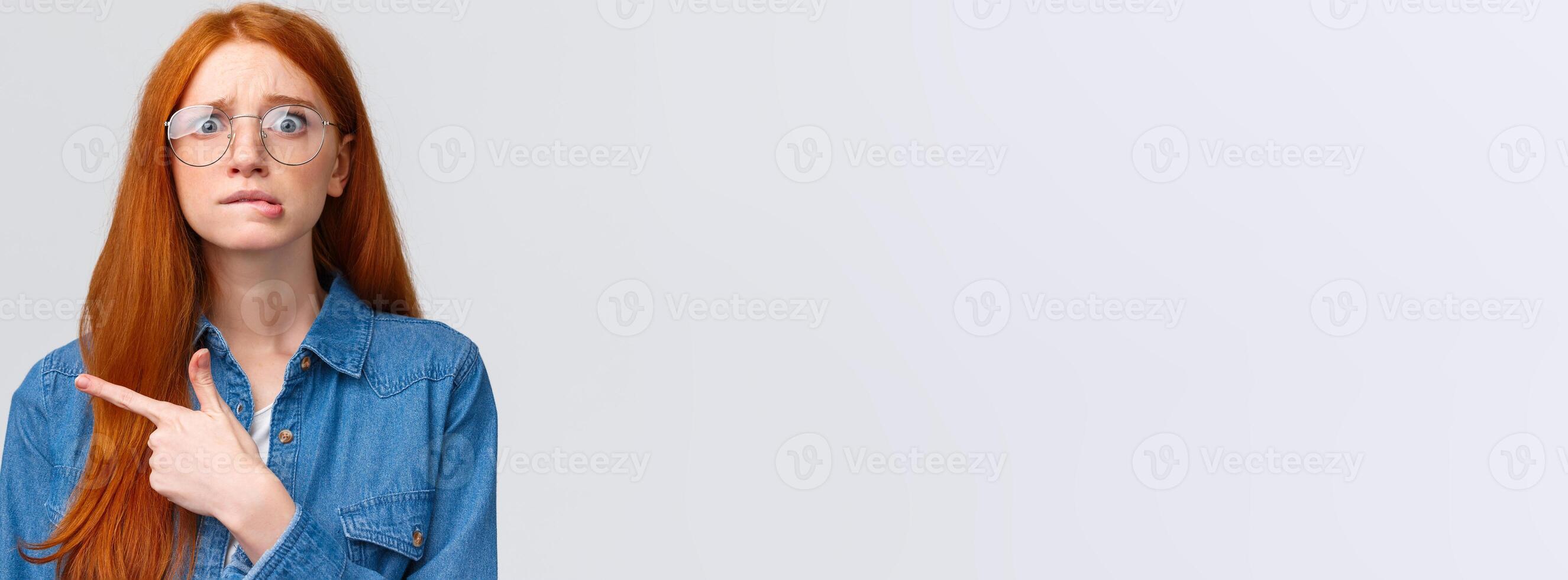 Nervousness, people and education concept. Anxious timid and insecure cute redhead girl in glasses, student worrying biting lip, hesitating telling something, pointing finger left, white background photo