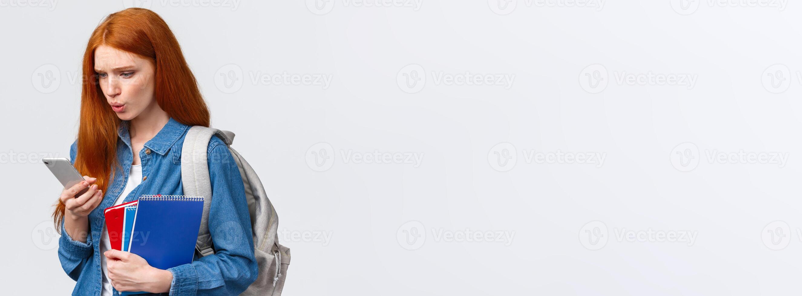 What is that. Confused and displesaed. intense troubled redhead teenage, college girl with backpack and notebooks walking university and reading strange text on smartphone, white background photo