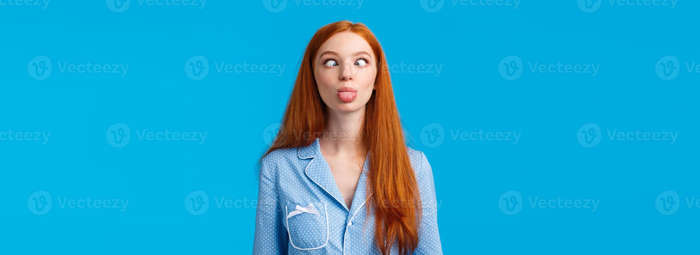 Be funny and silly. Redhead carefree and enthusiastic caucasian girl squinting, making goofy expression, showing tongue, standing blue background in nightwear, fool around photo