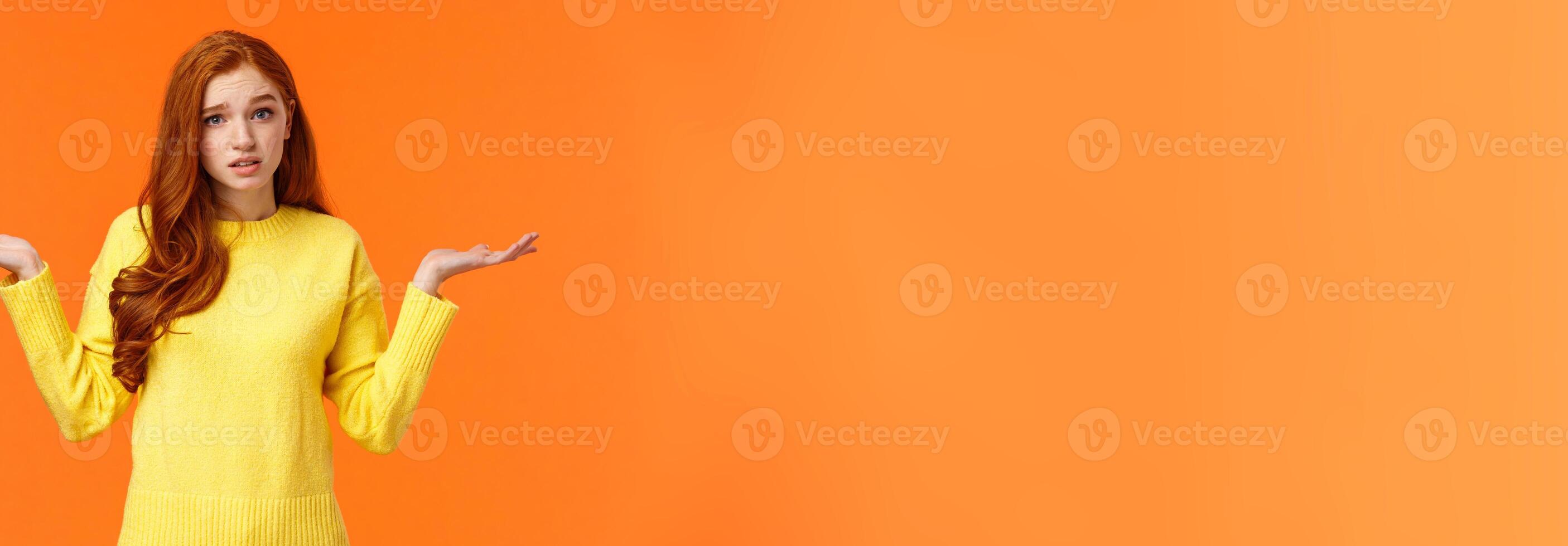 Perplexed cute redhead girl, shrugging with raised arms and looking confused, dont know answer, cant help, feeling confused or indecisive, cannot understand, standing orange background photo
