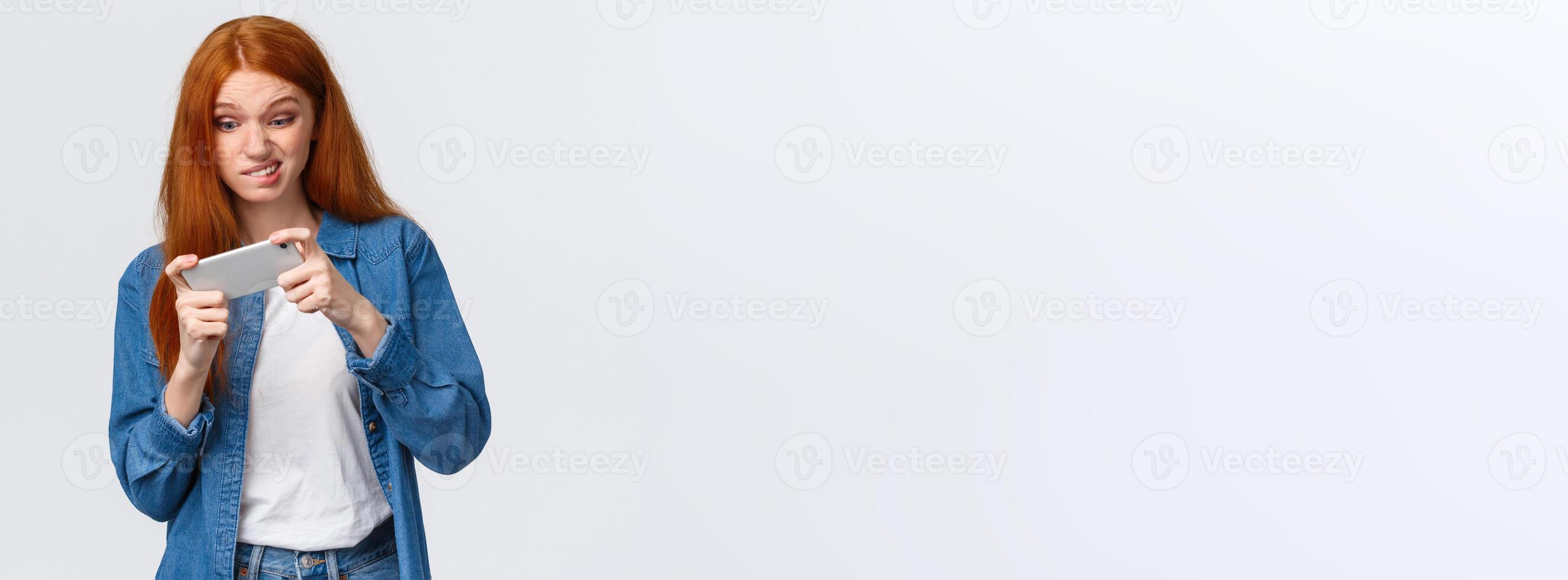 Girl having intense fighting round in game, tap screen and trying beat friends score in app. Excited good-looking redhead female student wasting time playing smartphone arcade, white background photo