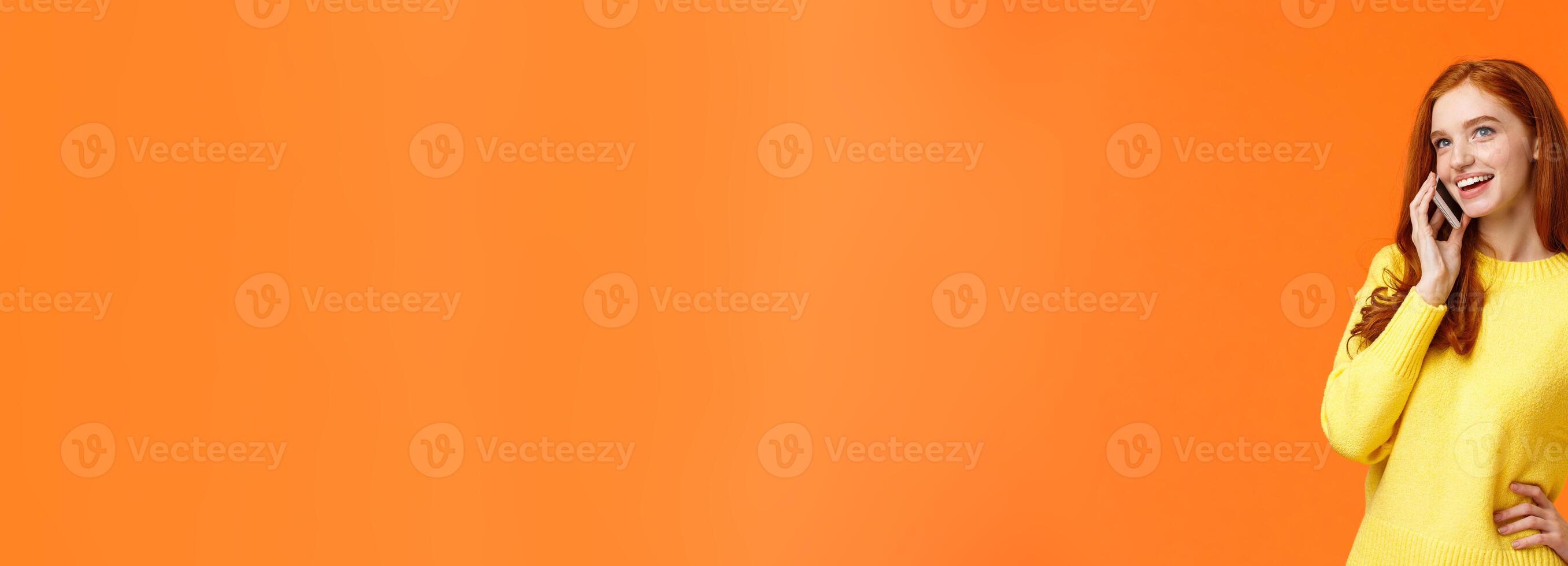 Outgoing pretty redhead girl having conversation on phone, calling friend, making arrangements, woman booking hotel or trip using mobile, speaking via smartphone, standing orange background photo