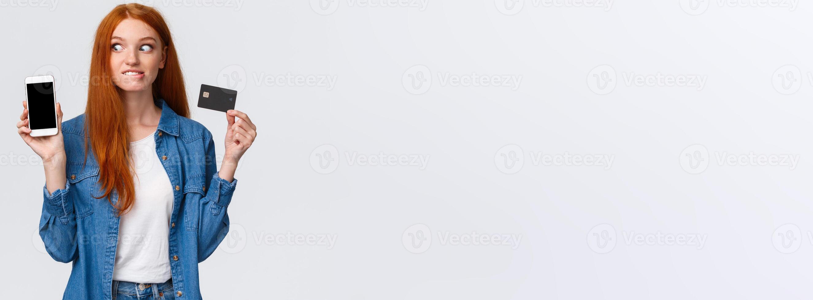 Shopaholic cant hold herself want buy something online. Eager and excited redhead girl see tempting price internet store, biting lip thrilled stare smartphone display, hold credit card photo