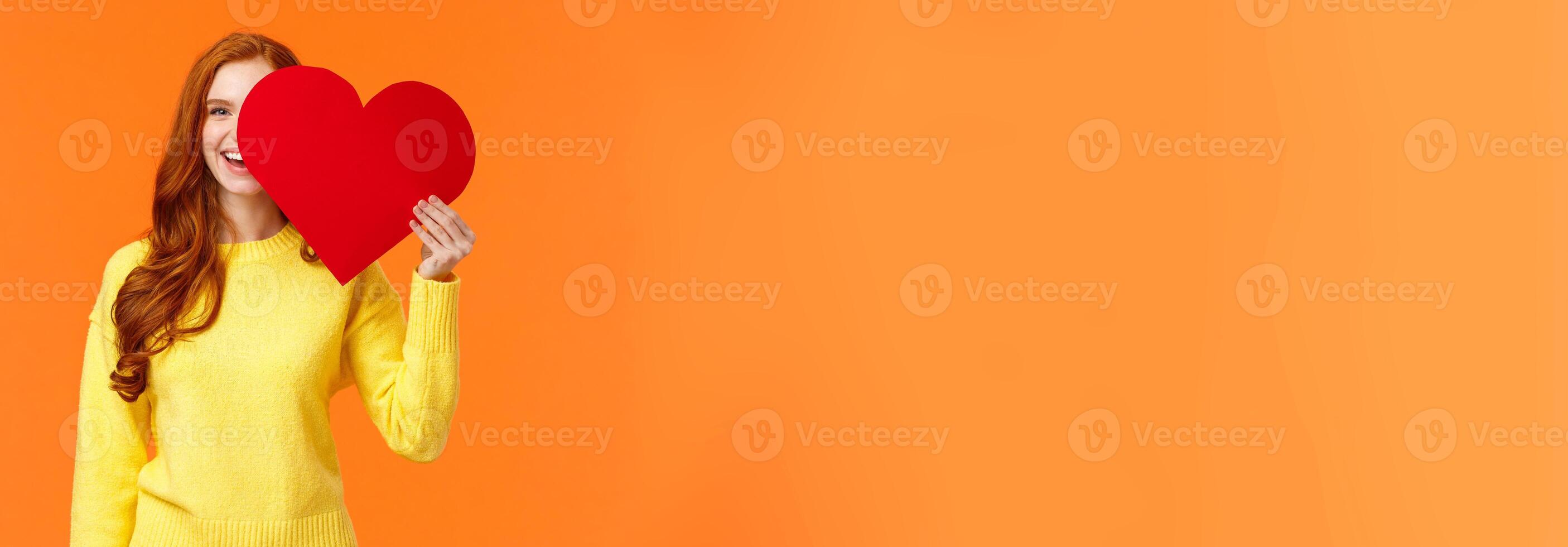 Holidays, romance concept. Cheerful girl showing affection, give heart to girlfriend, hiding half face behind valentines card, smiling joyfully, prepare romantic gesture for date, orange background photo