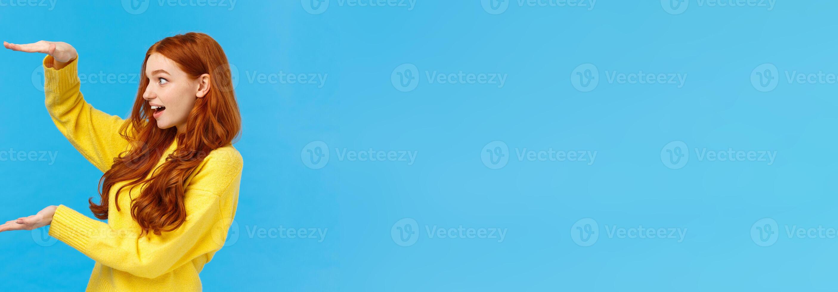 Impressed and startled excited cute redhead female receive big gift, shaping large object or product over copy space, holding hands as if carry enourmous present, smiling astonished, blue background photo